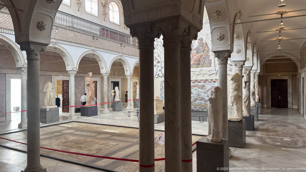 Columned Bardo Museum hall with classical statues, wall mosaics, arches, red barriers, and patterned floors.
