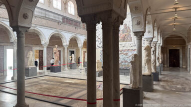 Columned Bardo Museum hall with classical statues, wall mosaics, arches, red barriers, and patterned floors.