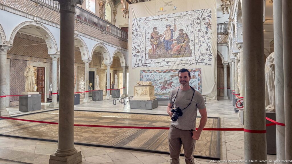 Man holding a camera standing in a large gallery with mosaics, statues, columns, and arched palace architecture.