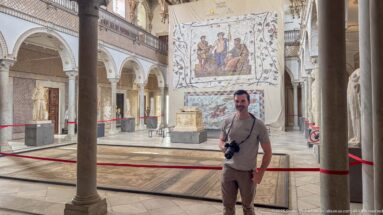 Man holding a camera standing in a large gallery with mosaics, statues, columns, and arched palace architecture.