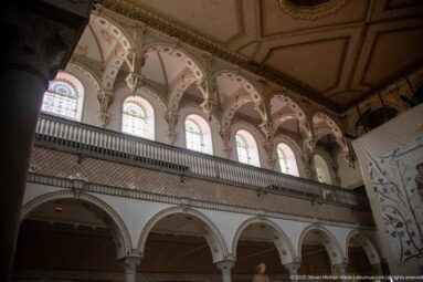 Upper gallery arcade with arched windows, decorative spandrels, lattice balustrade, and columns below.