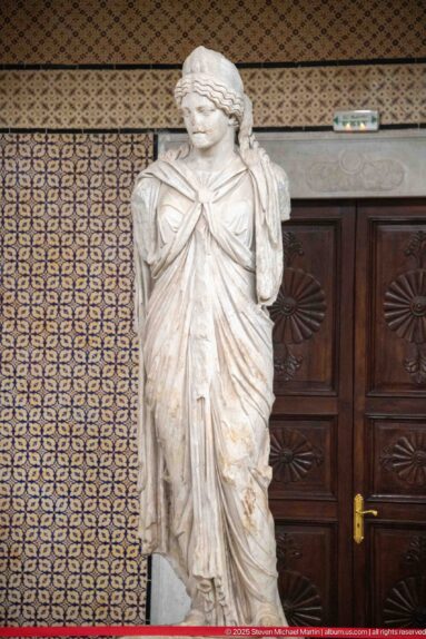 Standing marble woman wrapped in a heavy mantle with layered folds and head turned slightly to one side.