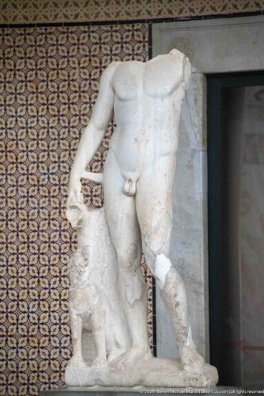 Headless nude marble male statue standing beside a tree-trunk support, with missing arm and lower leg.