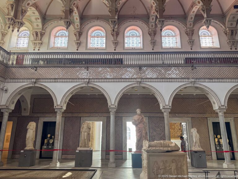 Arcaded Bardo Museum hall with classical statues below a row of upper arched windows and decorative panels.