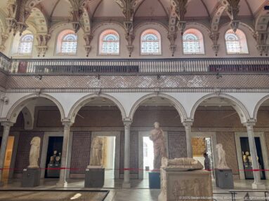 Arcaded Bardo Museum hall with classical statues below a row of upper arched windows and decorative panels.