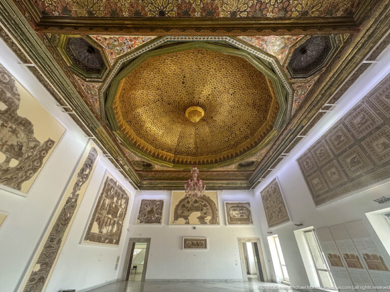 Gallery with an ornate octagonal gilded ceiling, central chandelier, and mosaics displayed on white walls.
