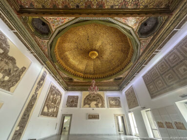 Gallery with an ornate octagonal gilded ceiling, central chandelier, and mosaics displayed on white walls.
