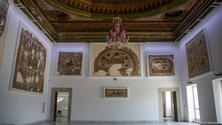 Gallery wall with several large mosaics beneath an ornate ceiling and pink chandelier.