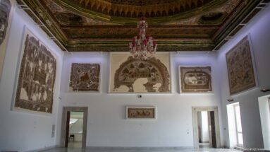 Gallery wall with several large mosaics beneath an ornate ceiling and pink chandelier.