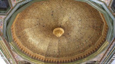 Close upward view of an octagonal decorated dome with dense interlaced ornament and a central hanging light.