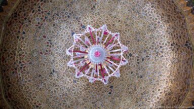 Pink-and-clear glass chandelier seen from below against a densely patterned golden ceiling.