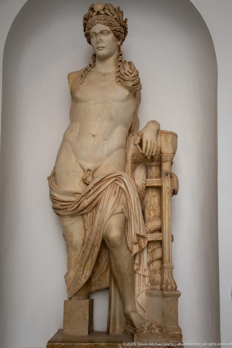 Seated marble male figure with bare torso, leafy wreath, draped mantle, and carved support beside one arm.