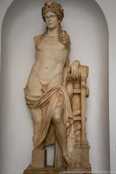 Seated marble male figure with bare torso, leafy wreath, draped mantle, and carved support beside one arm.