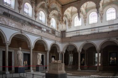 Two-level Bardo Museum gallery with arches, columns, upper windows, balustrades, and classical sculptures displayed around the floor.