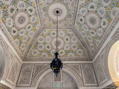 Polychrome palace ceiling with floral rosettes, geometric panels, shallow vaults, and a dark multicolored hanging glass lantern.
