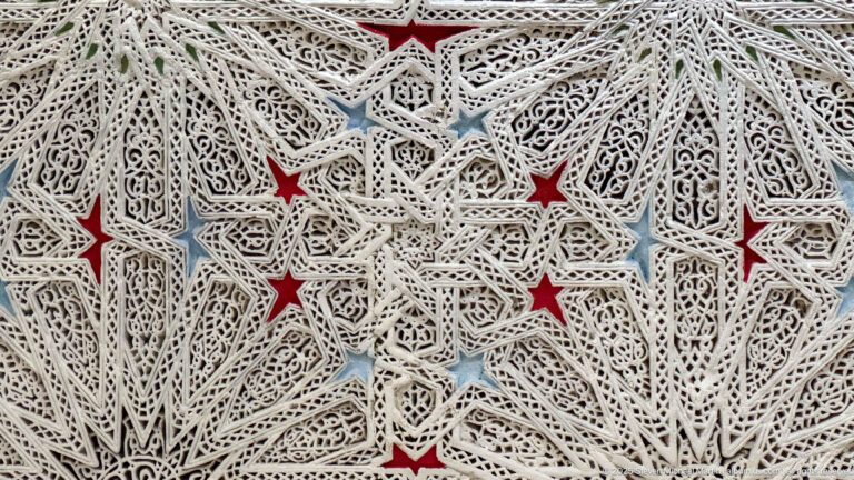 Cream openwork plaster lattice with interlaced stars, pierced arabesque carving, and small red and blue painted recesses.