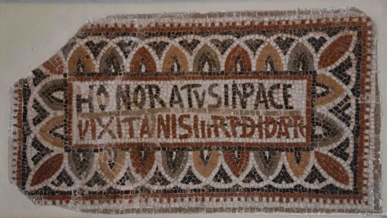 Rectangular Christian mosaic inscription framed by repeating red, black, and cream pointed leaf-shaped motifs.