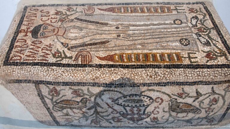 Angled view of a mosaic-covered funerary box or tomb with Latin inscriptions, birds, vessels, and vegetal motifs.