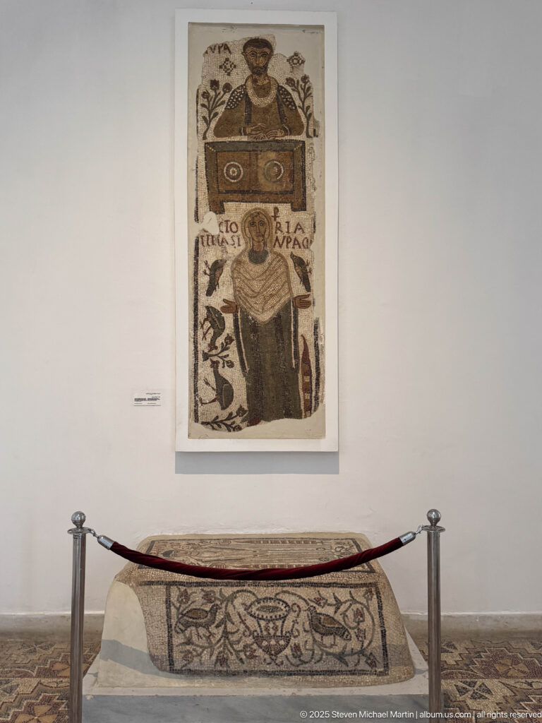Tall Christian portrait mosaic mounted above a mosaic-covered funerary monument behind gallery barriers.