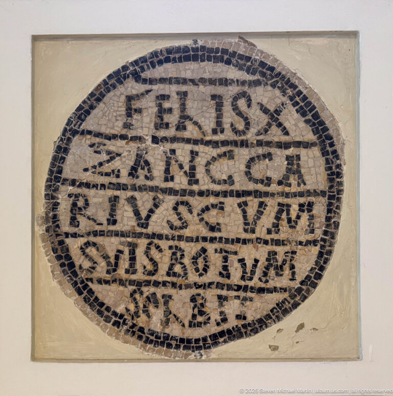 Oval Christian mosaic containing multiple lines of Latin text inside a thick dark tessellated border.