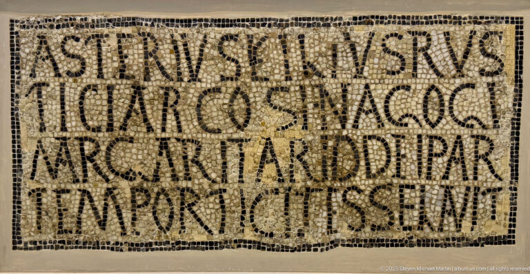 Rectangular Christian mosaic filled with several lines of large Latin capital letters inside a narrow dark border.