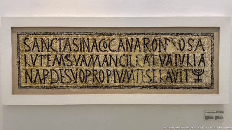 Long rectangular Christian mosaic inscription with several lines of Latin text and a small cross-like symbol at lower right.