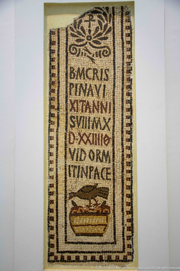 Narrow Christian funerary mosaic with Latin inscription, floral or cross-like ornament, and a small reclining figure at the bottom.
