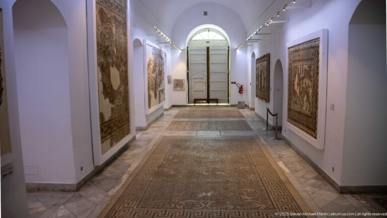 Long vaulted Bardo Museum corridor with mosaic floor pavements, wall-mounted panels, and a tall arched doorway at the far end.