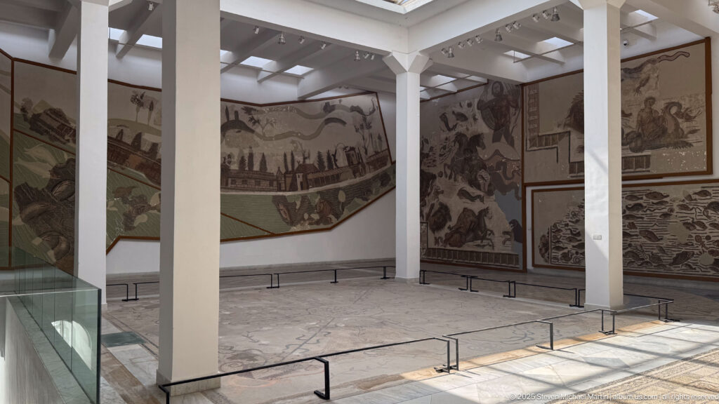 Spacious white-columned gallery lined with large Roman narrative mosaics and low protective barriers.