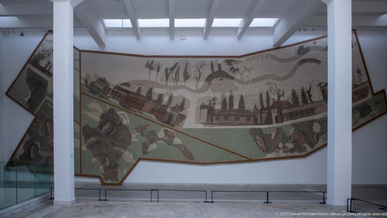Large irregular Roman mosaic with buildings, trees, people, animals, and broad landscape bands mounted on a gallery wall.