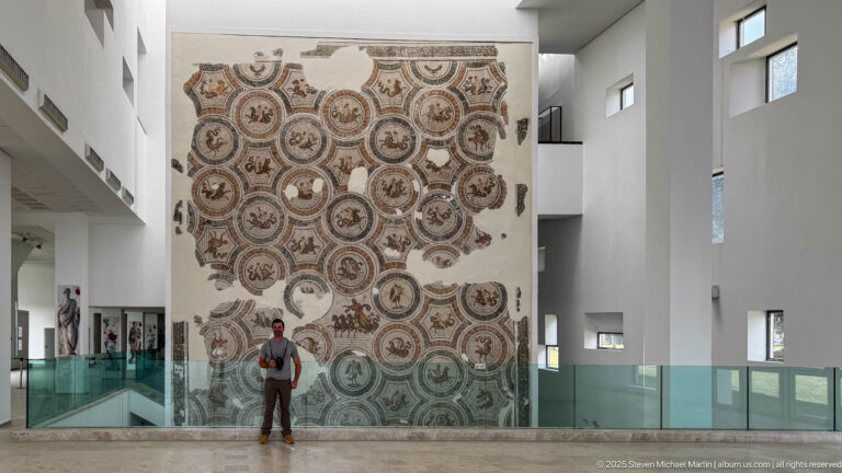 Large wall-mounted Roman mosaic of linked figural medallions with a visitor standing below for scale.