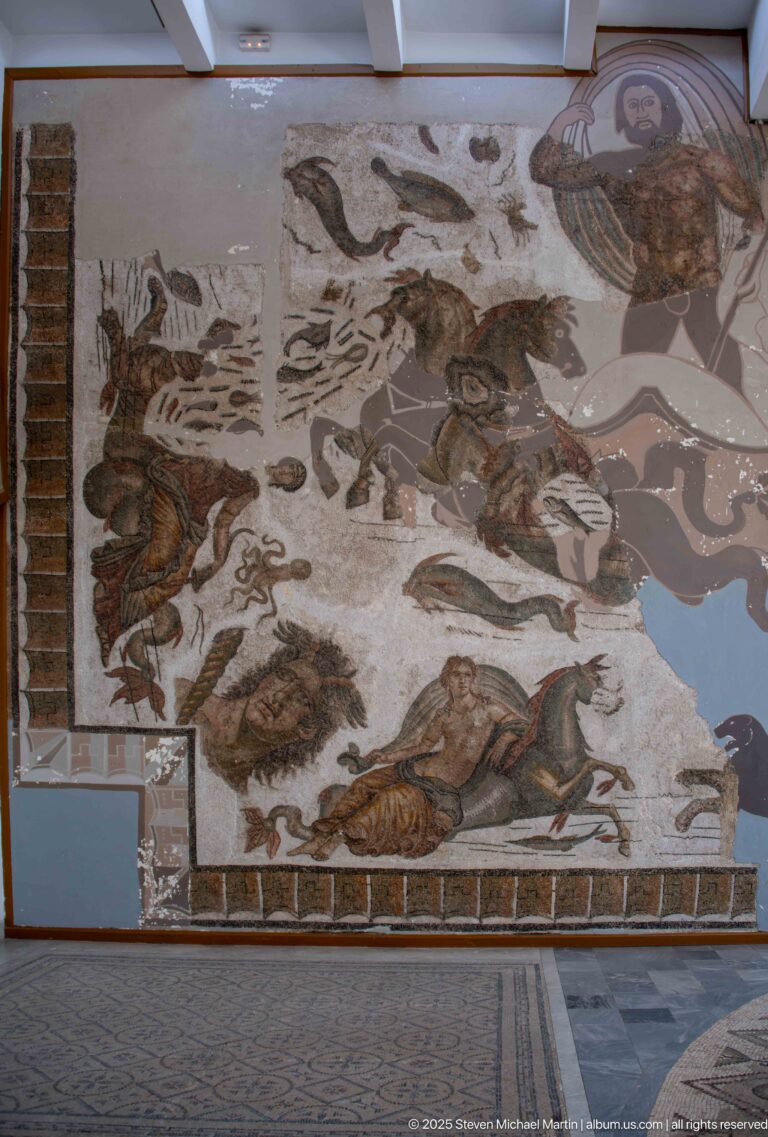 Large wall-mounted Roman mosaic with wild animals scattered across a pale landscape and irregular areas of loss.