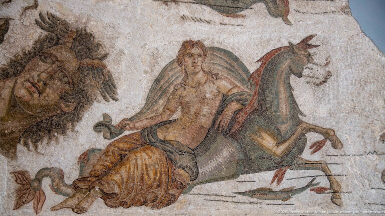 Mounted man on a horse beside a large lion in a Roman mosaic, with detailed mane, harness, and colored tesserae.