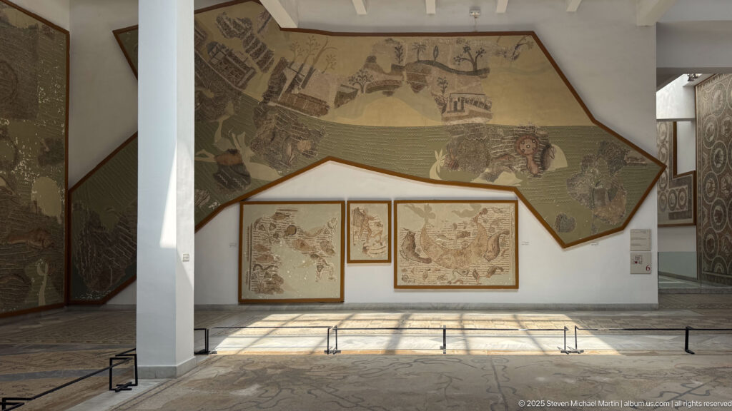 Sunlit museum gallery with a monumental irregular Roman narrative mosaic above three smaller framed panels.