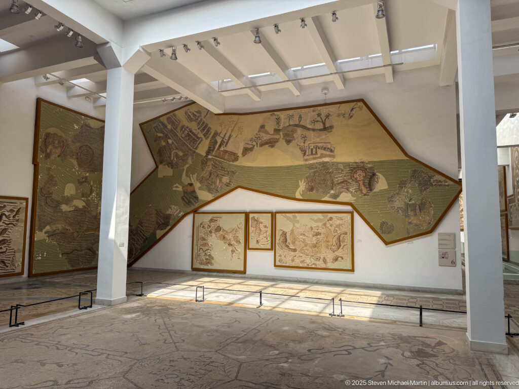 Wide Bardo Museum gallery with a monumental wall mosaic, smaller framed panels, columns, and mosaic pavements underfoot.
