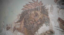 Close detail of a lion's head with curling mane rendered in dark and ochre tesserae on a damaged Roman mosaic.