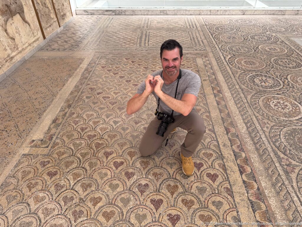 Visitor kneeling on a protected gallery route beside extensive floor mosaics with scalloped, bordered, and geometric patterns.