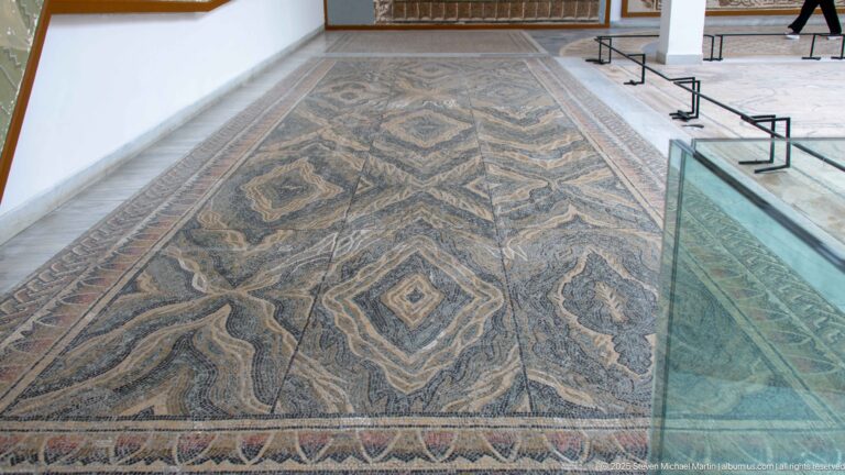Large rectangular floor mosaic with nested diamonds, angular ribbon patterns, vegetal motifs, and a patterned outer border.