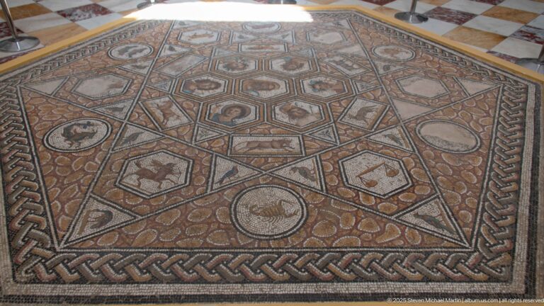 Square Roman floor mosaic divided into geometric compartments with portrait heads, animals, and motifs inside a braided border.