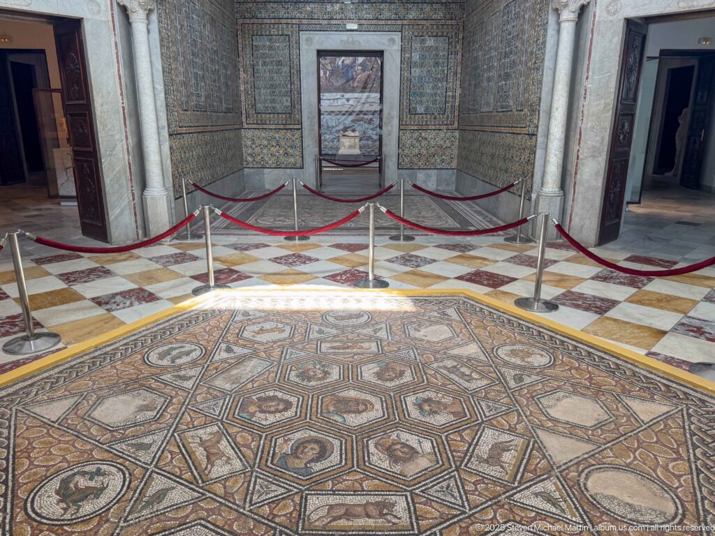 Large geometric and figural floor mosaic in an ornate gallery with mosaic-covered walls, marble columns, and red barriers.