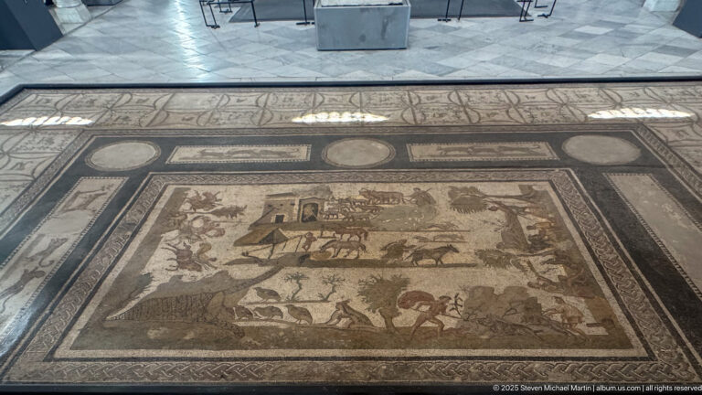 Large Roman floor mosaic with villa buildings, cart, people, and wild animals inside broad geometric framing panels.