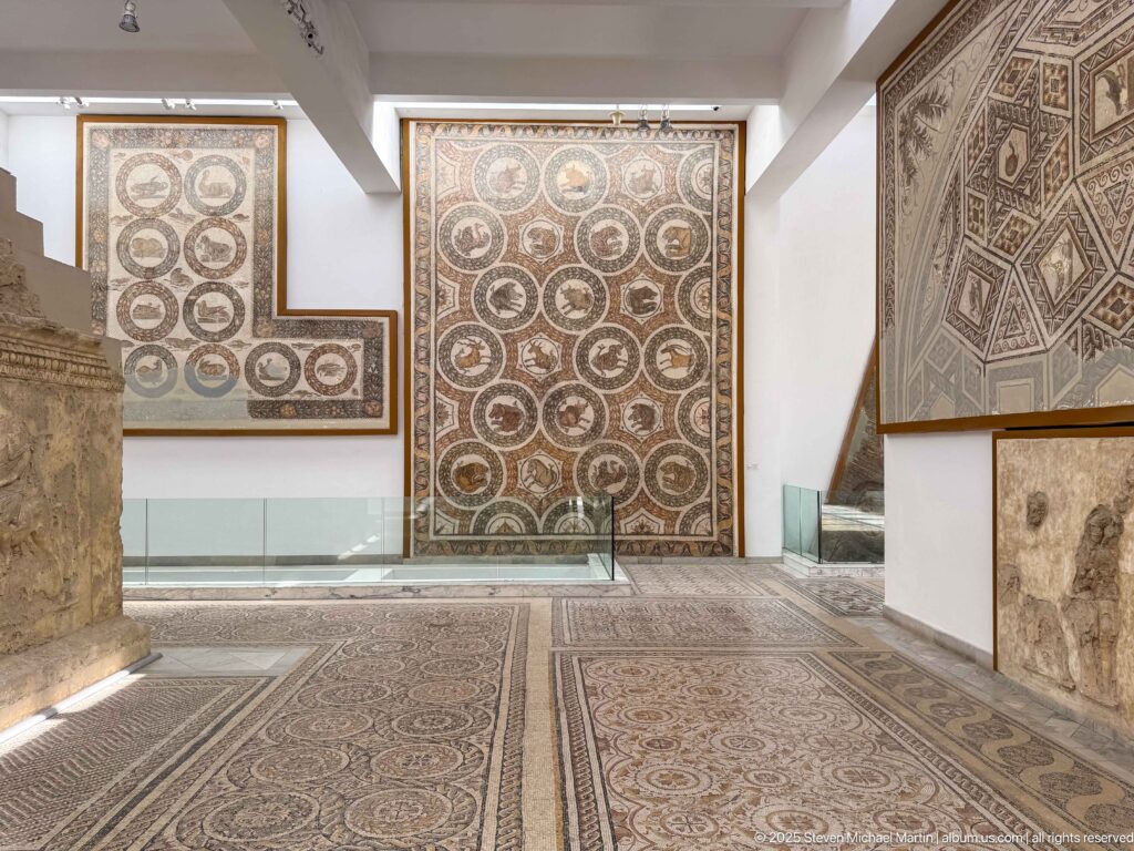 Bright museum gallery with monumental Roman mosaics mounted on walls and patterned mosaics covering parts of the floor.