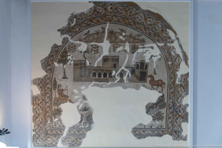 Arched Roman mosaic fragment with villa buildings, tall trees, birds, animals, ornate border, and extensive central losses.