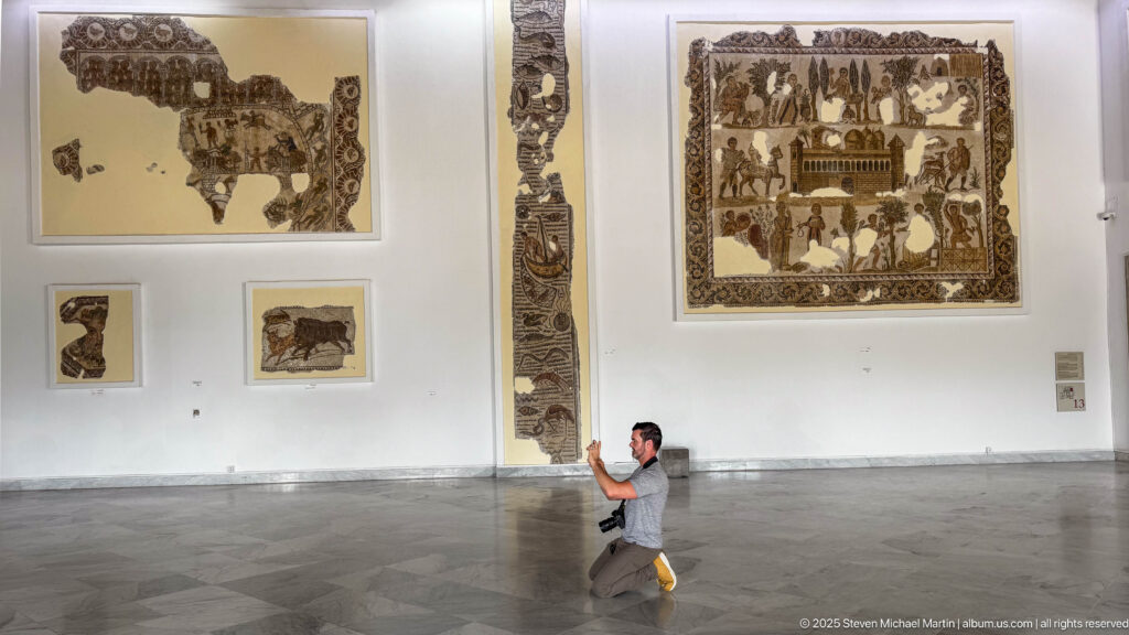 Visitor walking through a white museum gallery displaying large narrative mosaics, animal fragments, and a tall vertical mosaic panel.