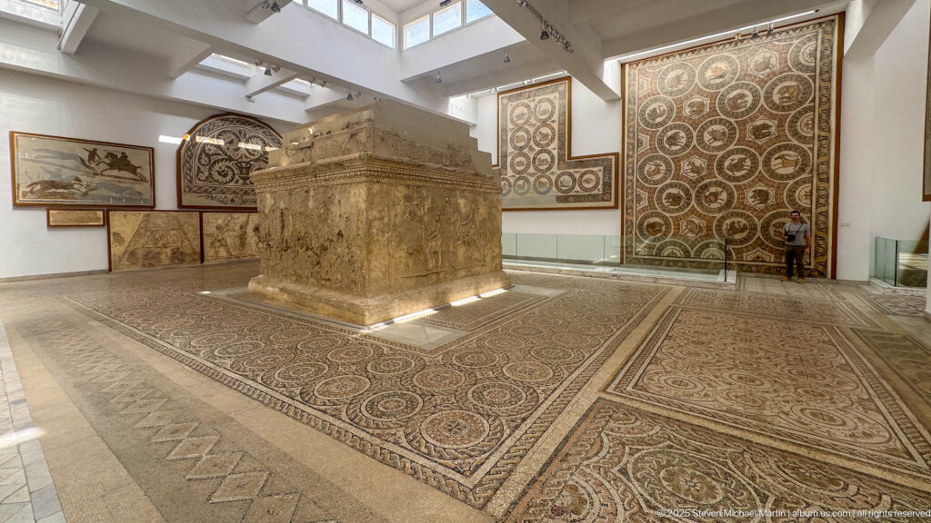 Large rectangular carved stone monument standing on a mosaic floor amid wall-mounted Roman mosaics.