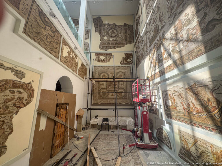 High museum room with wall-mounted mosaics, scaffolding, equipment, stored materials, and strong sunlight across one wall.