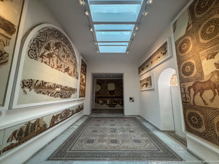 Long skylit Bardo Museum gallery with a large geometric floor mosaic and monumental wall-mounted mosaics on both sides.