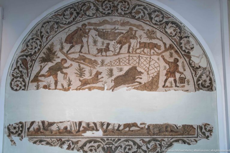 Semicircular Roman hunting mosaic fragment with hunters, game animals, dogs, trees, and a large net above a broad missing band.