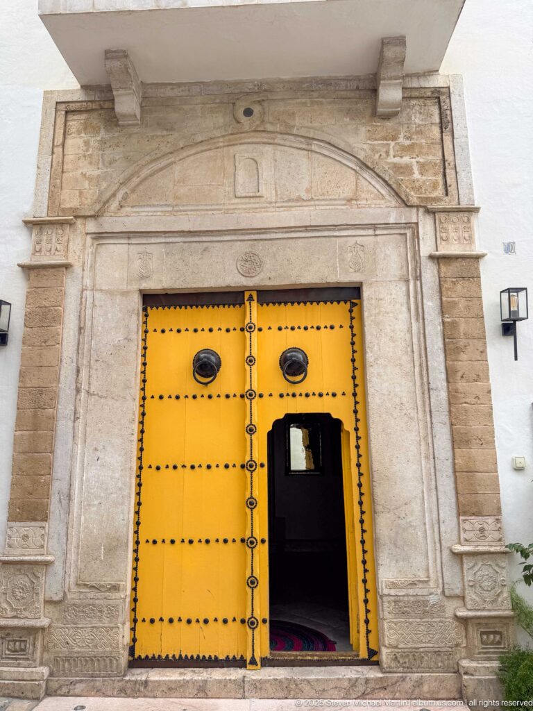 Large yellow studded double door with inset wicket door, framed by a carved pale stone portal at Dar Jeld Hotel.