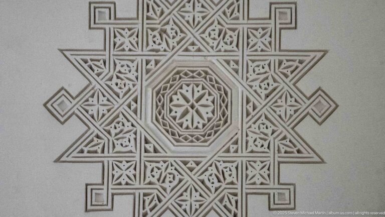 Pale carved plaster panel with central star, nested octagons, pointed projections, and angular interlaced bands.